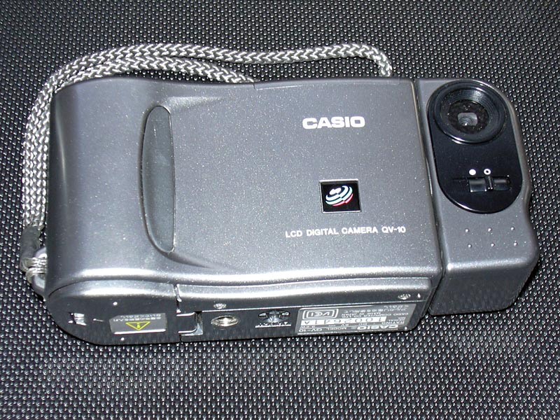 La cámara Casio QV-10 de 1995, gris, con el objetivo montado en un cuerpo giratorio a la derecha y el rótulo LCD DIGITAL CAMERA QV-10 impreso en el frontal.